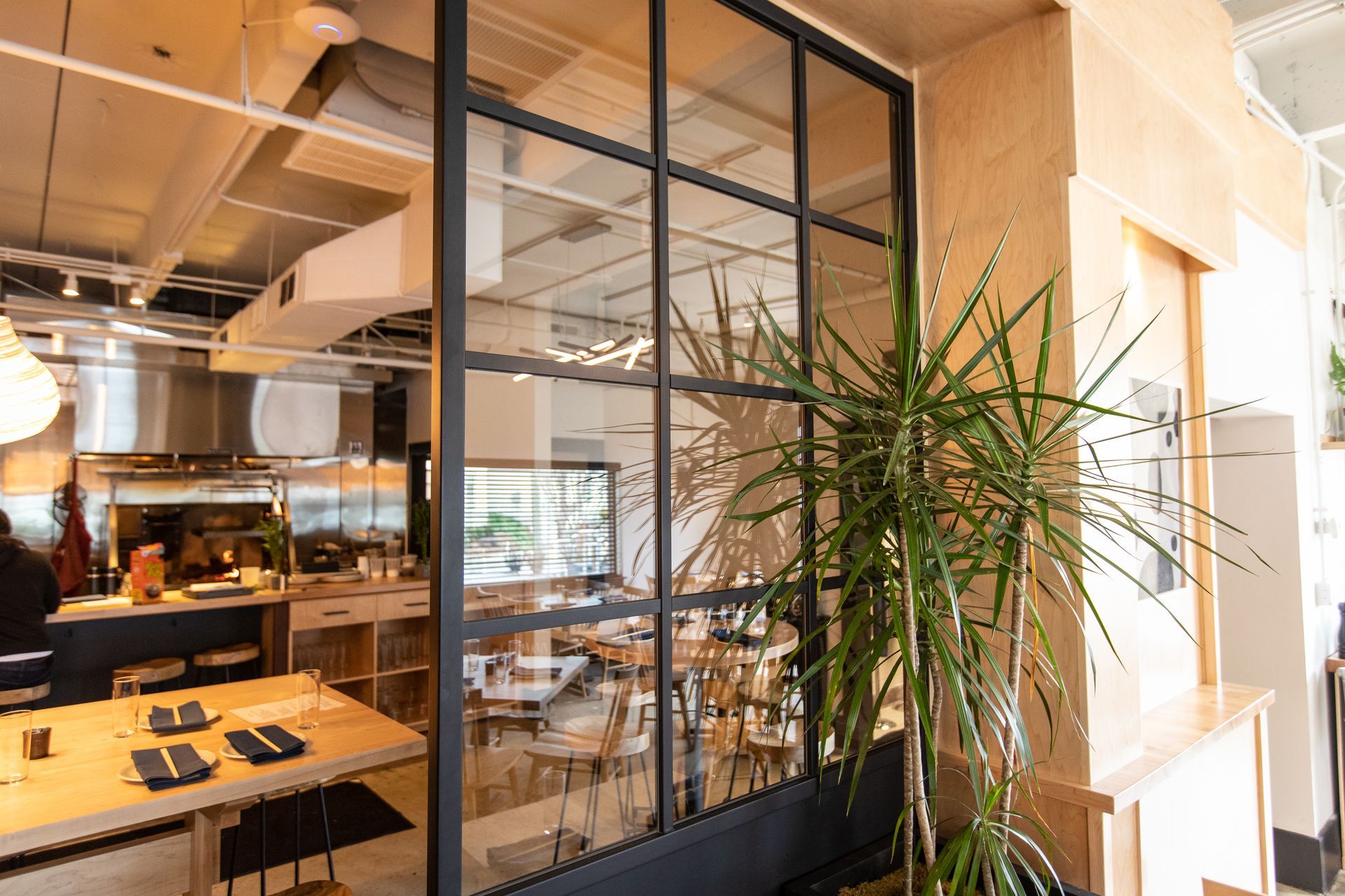Restaurant dining room with an open kitchen behind a steel-framed glass partition and pale timber joinery
